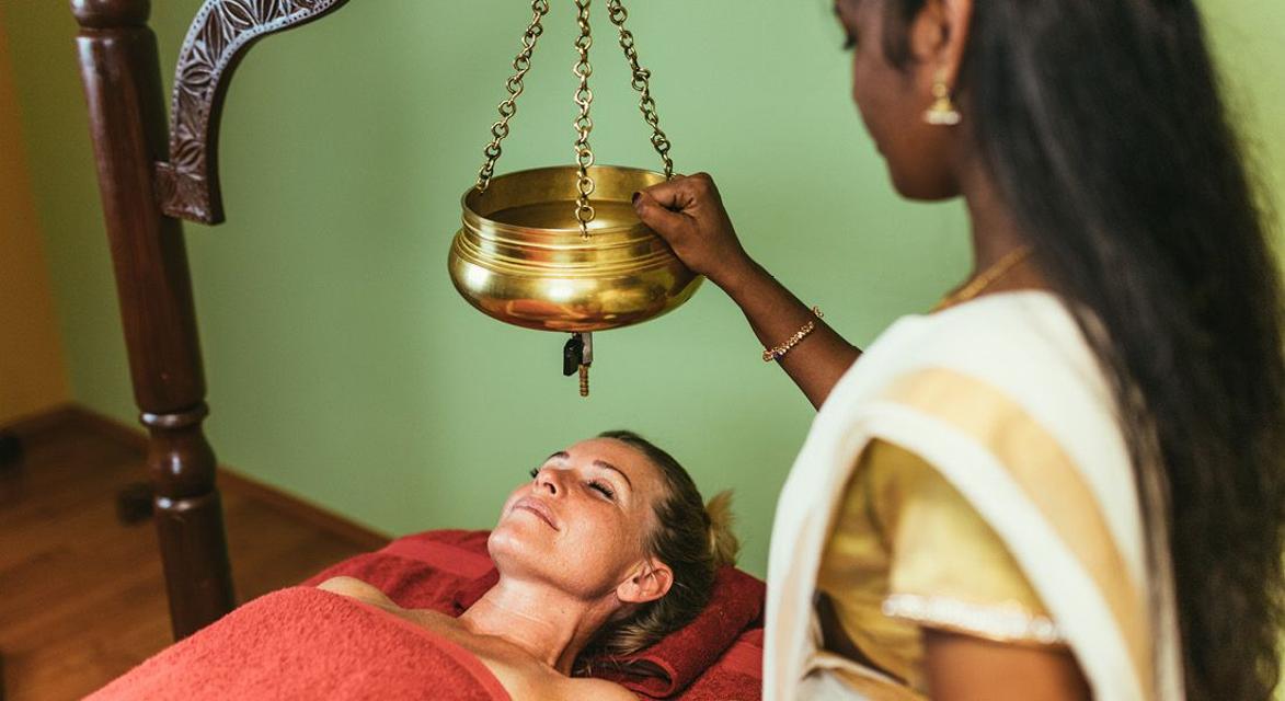 Auf dem Bild sehen Sie eine Frau bei einer ayurvedischen Therapieanwendung - hier fließt ein feiner Strahl aus warmen Öl auf ihre Stirn.