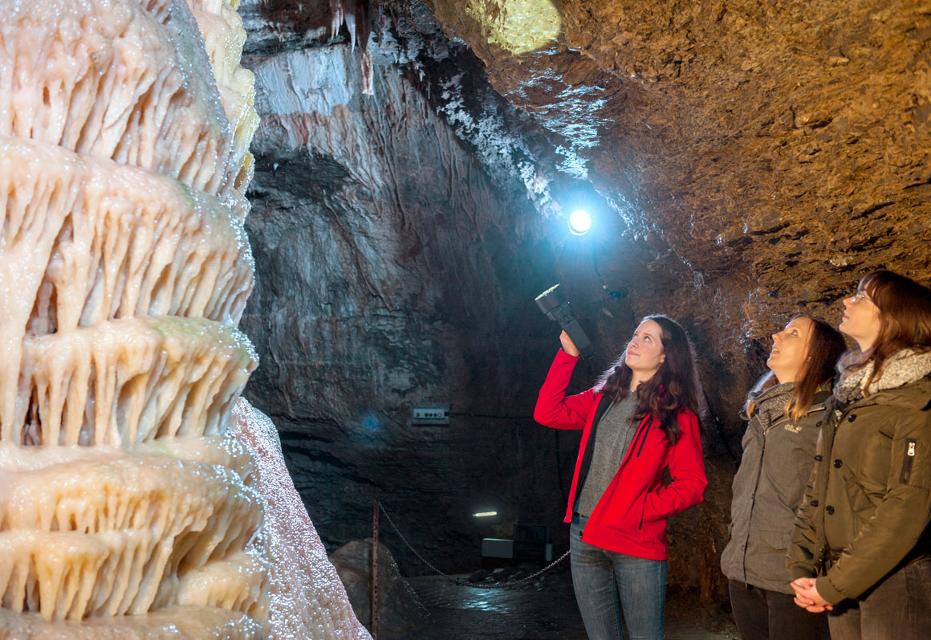 Drei Personen nehmen in einer Tropfsteinhöhle an einer Höhlenführung teil. Die Gästeführerin zeigt mit der Taschenlampe auf einen großen Tropfstein mit dem Namen Hochzeitstorte.