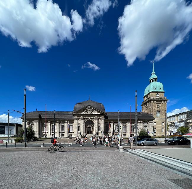 Blick vom Friedensplatz auf das Hessische Landesmuseum. Großer, langgestreckter Bau mit Portal in der Mitte. Rechts am Gebäude ein Turm mit türkisfarbenem Kupferblechdach, gewölbt mit Spitze.