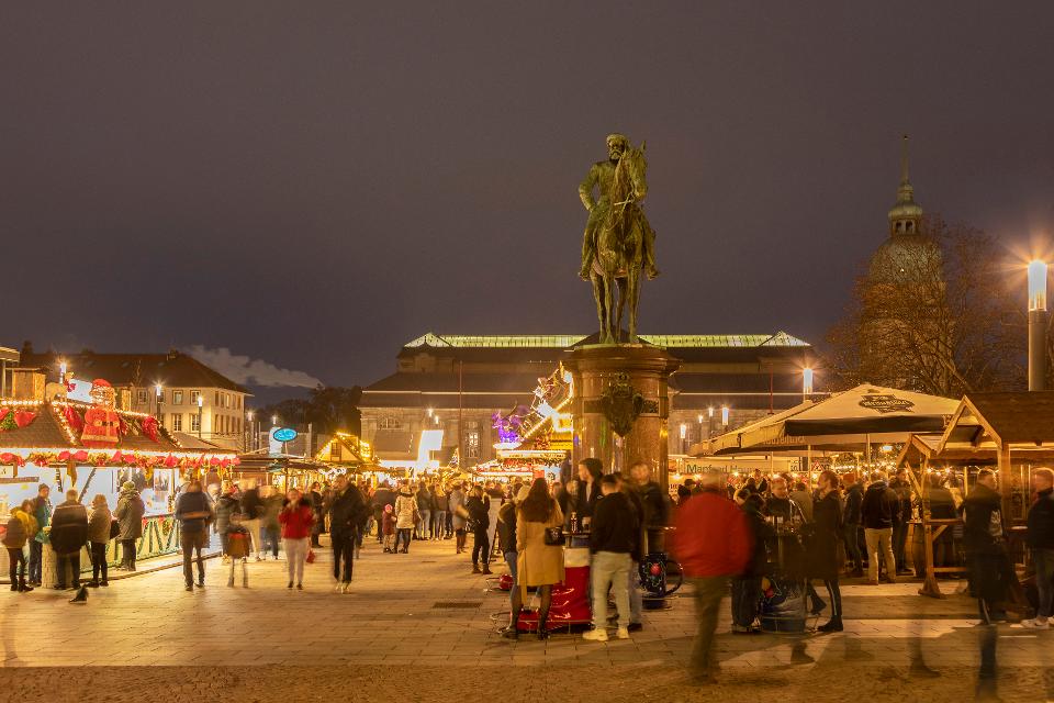 Ein beleuchteter Platz mit Weihnachtsmarktbuden, vielen Weihnachtsmarktbesucherinnen, im Vordergrund ein Denkmal mit einer Reiterfigur, im Hintergrund ein großes Gebäude mit einem Turm an der rechten Seite.