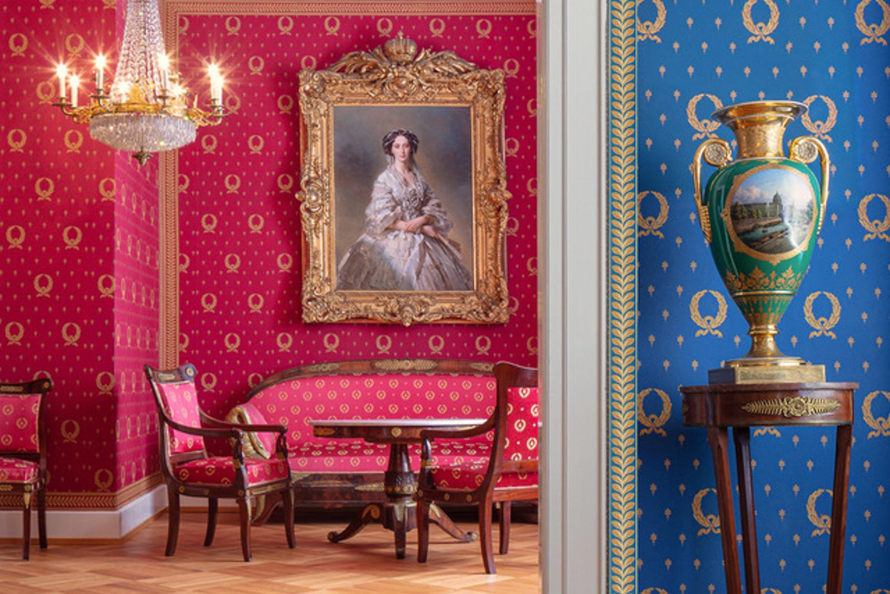 Blick durch einen Türrahmen, rechts blaue  Tapete mit goldenem Muster, davon eine hohe Vase auf einem schmalen Tisch. Im Raum dahinter eine Sitzgruppe aus Sofa und zwei  Stühlen, an der Wand rote Tapete und historisches Portrait einer Dame.