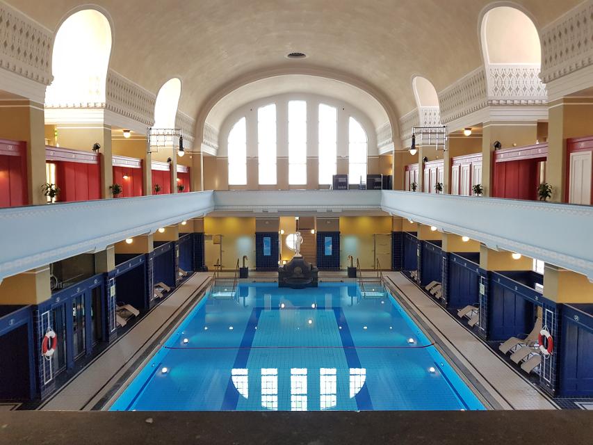 Blick von der Galerie auf das Schwimmbecken, links und rechts auf der Galerie sind die historischen Umkleidekabinen in roter Farbe zu sehen, das Becken leuchtet blau, die Säulen um das Becken sind mit dunkelblauen glänzenden Kacheln gefliest.