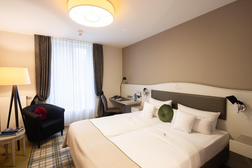 Zimmer mit Doppelbett, helle, warme Erdfarben, der Teppich ist kariert, in der Ecke steht eine Stehlampe und ein Sessel.