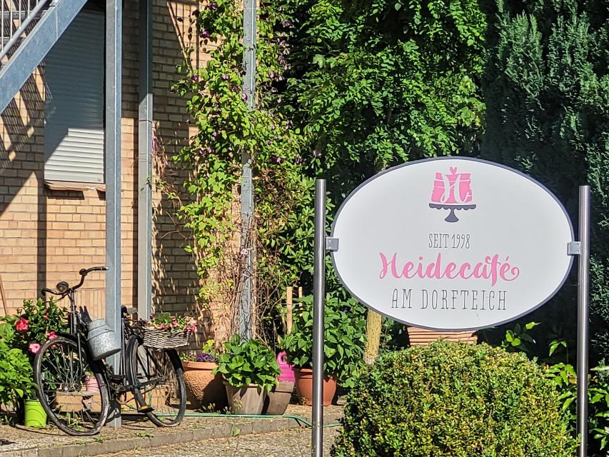 Auf einem ovalen, weißen Schild ist, in rosa Buchstaben, der Name des Cafès "Heidecafè am Dorfteich zu lesen. Drumherum sieht man grüne Büsche und Sträucher, ein altes Fahrrad und verschiedene Blumentöpfe als Dekoration.