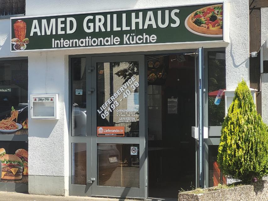 Zu sehen ist eine Glasfront mit grauem Rahmen. Darüber hängt das Schild der Gastronomie. Auf grünem Hintergrund steht in weißen Buchstaben "Amed Grillhaus" internationale Küche. Außerdem sind unterschiedliche Speisen, ein Dönerspies, eine Pizza, Burger und Spaghetti, zu sehen.