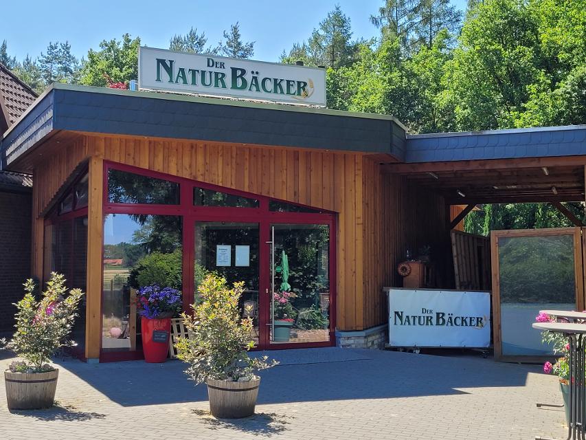 Auf einem, mit Holz verkleideten Ladengeschäft steht in großen, grünen Buchstaben der Name der Bäckerei "Der Naturbäcker". Davor stehen bunte Blumen und Sträucher in Kübeln. Im Hintergrund sieht man hohe Bäume.