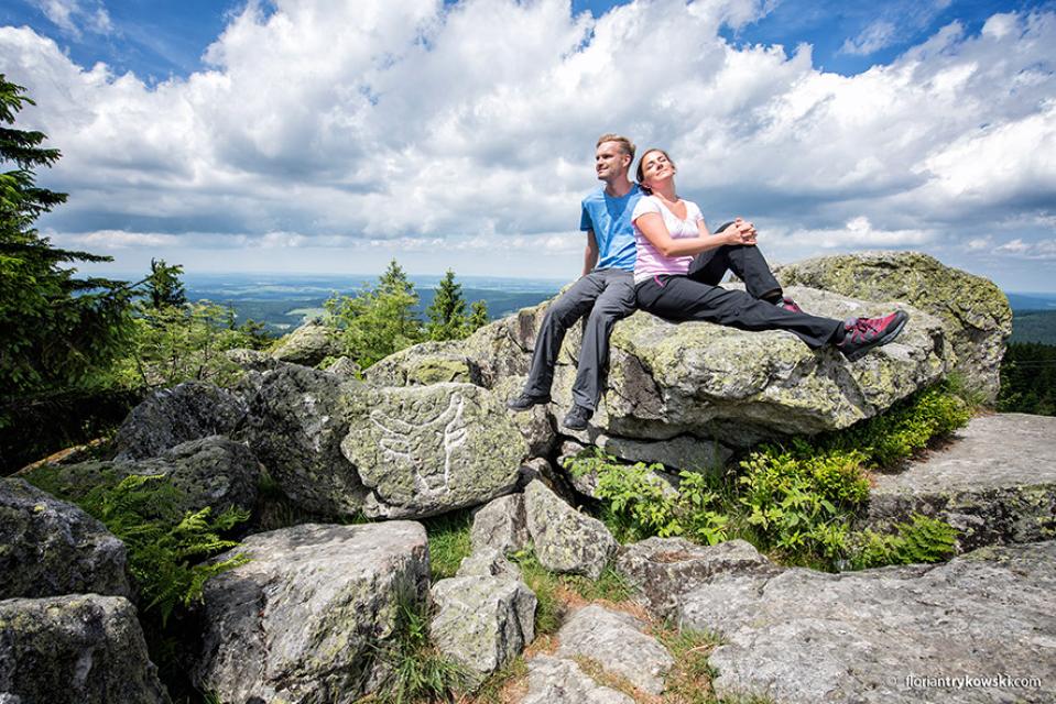 Zwei Wanderer sitzen auf dem Ochsenkopf-Gipfel auf einem Felsen.
                 title=