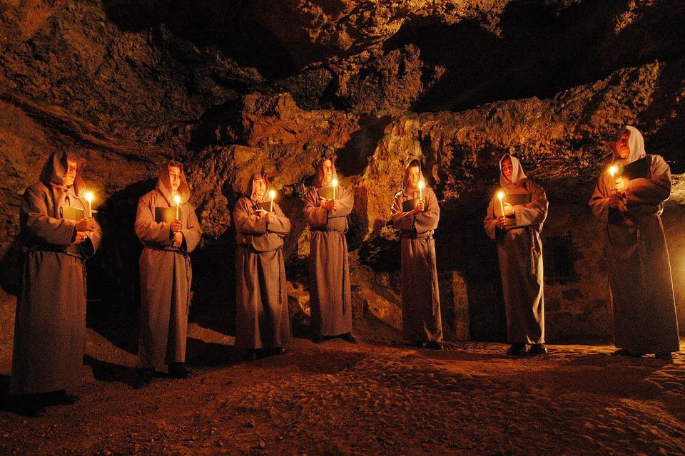 Sieben Männer in Mönchskleidung stehen in einer Reihe in einer Höhle. Sie haben Kerzen in der Hand. Die Höhle ist nur durch das Kerzenlicht beleuchtet. 