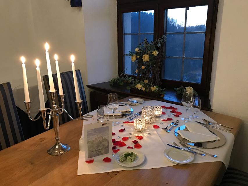 Ein Holztisch ist gedeckt mit weißer Tischdecke, Kerzen und Rosenblättern. Im Hintergrund ein Fenster.