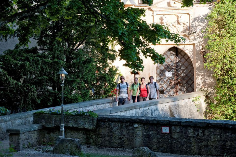 Vier Wanderer verlassen die Burg über die Brücke. Über dem Eingangstor erkannt man das prunkvolle Wappen in Stein gemeiselt.