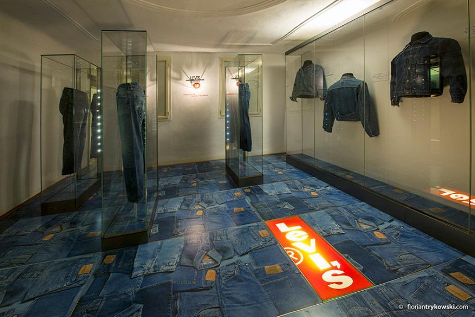 In einem Ausstellungsraum, dessen Boden das Motiv nebeneinanderliegender Jeans hat, hängen Jeans und Jeansjacken in Vitrinen.