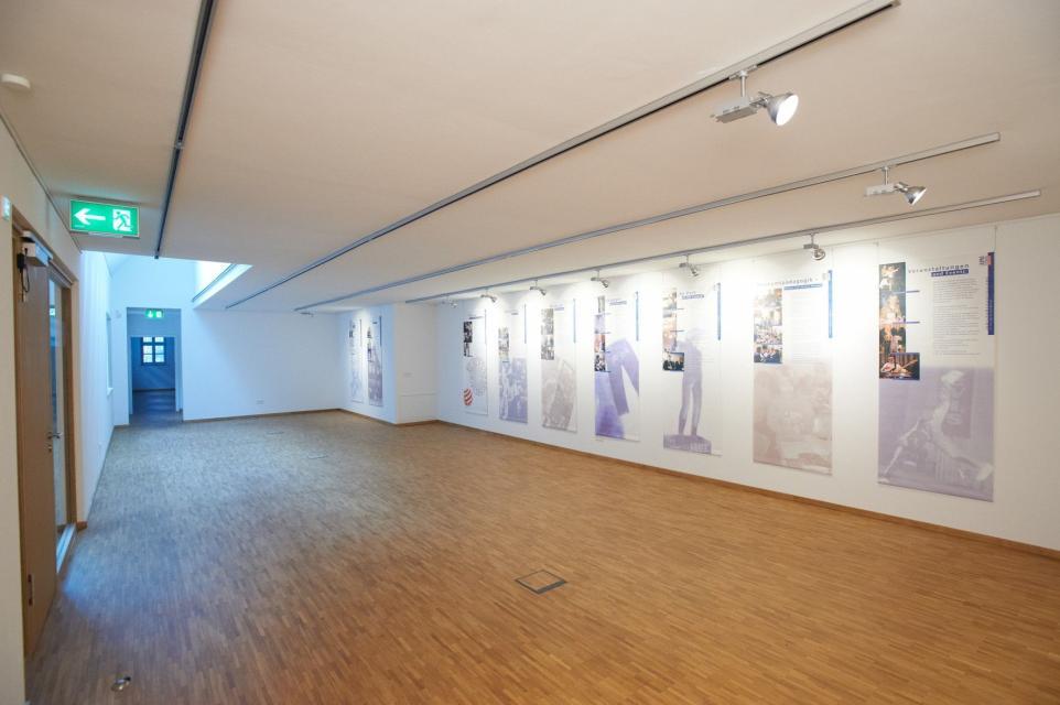 Ein großer, heller Ausstellungsraum mit Holzboden. An der Wand hängen verschiedene Informationstafeln mit Text und Bildern.