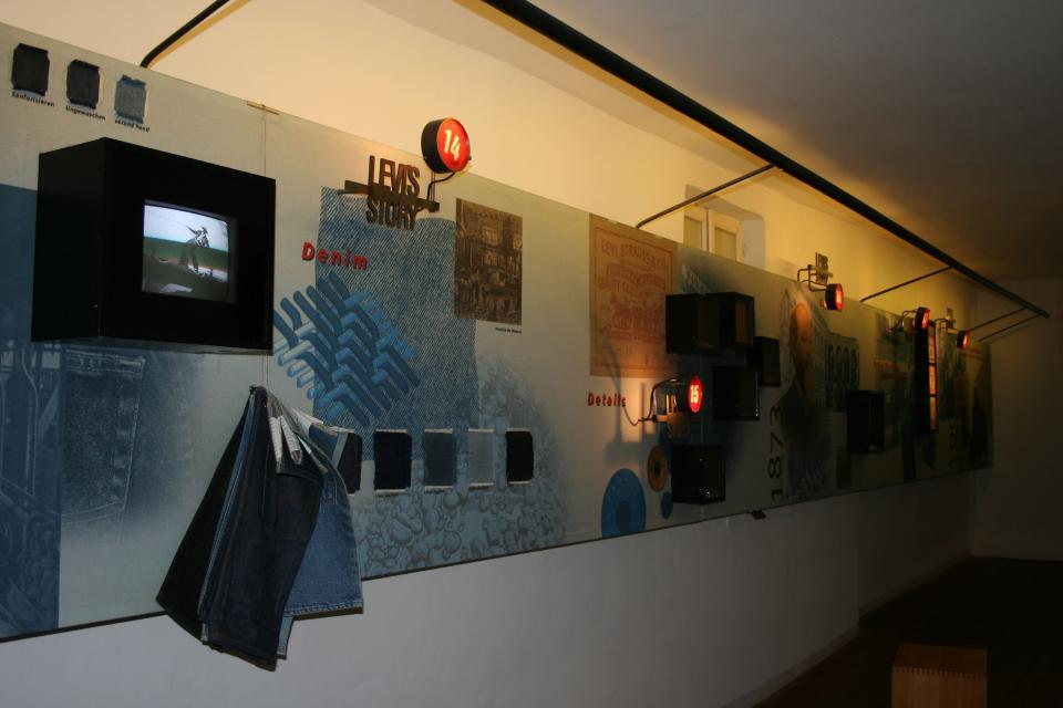 In einem Ausstellungsraum hängen an der Wand verschiedene Ausstellungsstücke, wie z. B. Jeansstoff. Zudem ist ein kleiner Monitor angebracht, auf dem ein Film zu sehen ist.