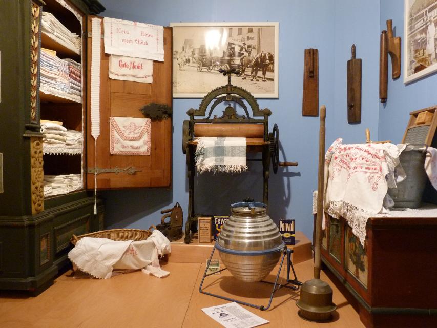 In einem blauen Zimmer steht links ein geöffneter Schrank, in dem Stoffe gestapelt sind. Im Raum befinden sich weitere Ausstellungsgegenstände.