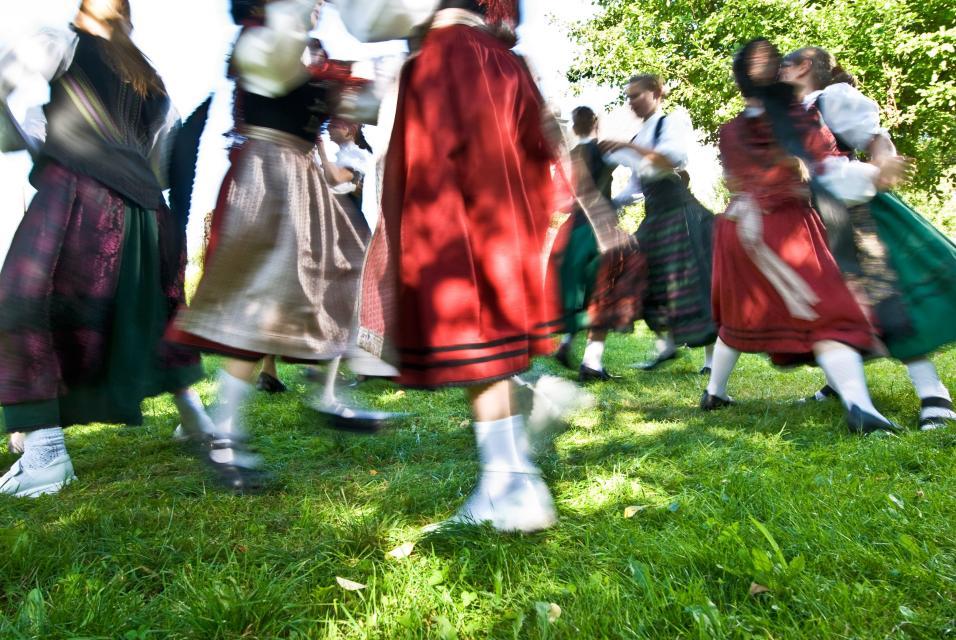 Mädchen in roter, grüner Tracht tanzen auf einer Wiese.