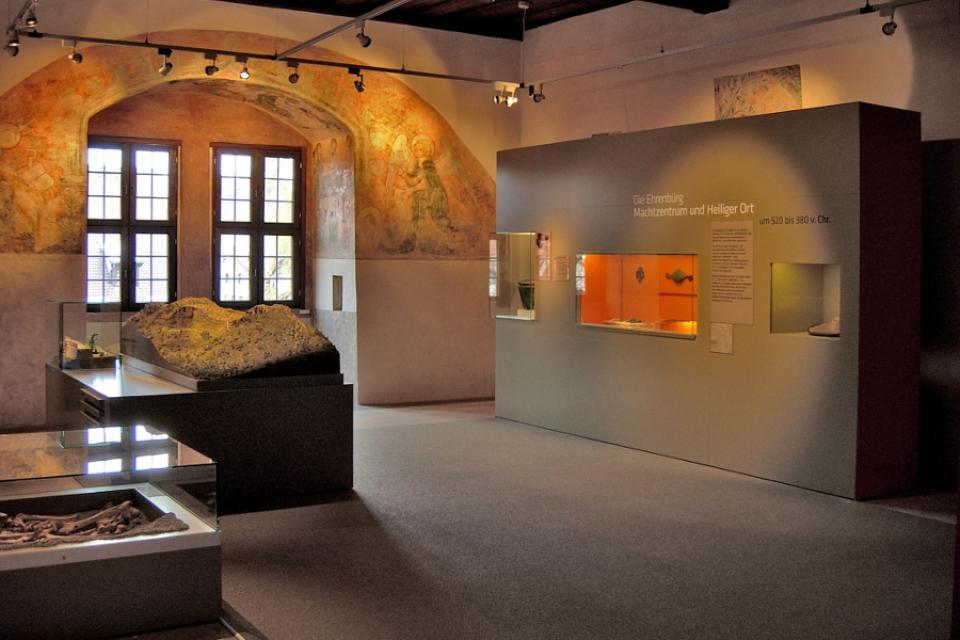 In einem Ausstellungsraum im Museum stehen zwei Glasvitrinen mit Exponaten. Vor einer Wand erhebt sich eine grauer Kasten, in dem weitere, kleine Vitrinen zu sehen sind.