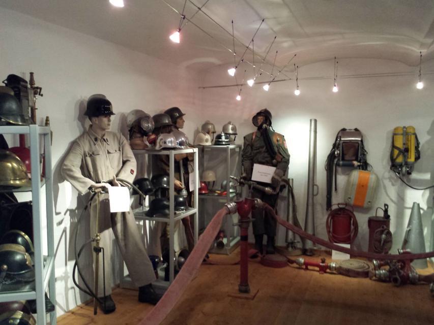 Ausstellungsraum mit zwei lebensgroßen Puppen in alter Feuerwehrmontur sowie weiteren Ausstellungsstücken in offenen Regalen.
                 title=