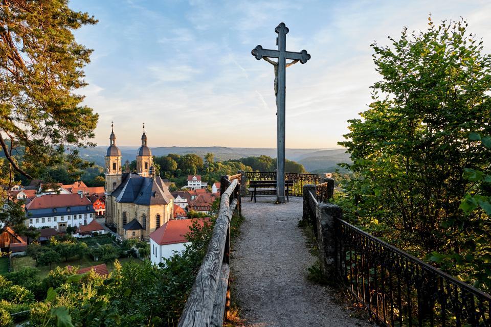 Ein Weg führt zu einem Aussichtspunkt mit Kreuz. Im Hintergrund ist eine Kirche, die Basilika im Sonnenuntergang.
