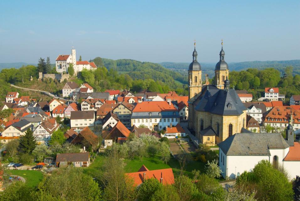 Ortseinsicht mit vielen Fachwerkhäusern, der Basilika und der Burg Gössweinstein zur Zeit der Kirschblüte. In der Ferne bilden grüne Wälder den Hintergrund zusammen mit blauem Himmel.