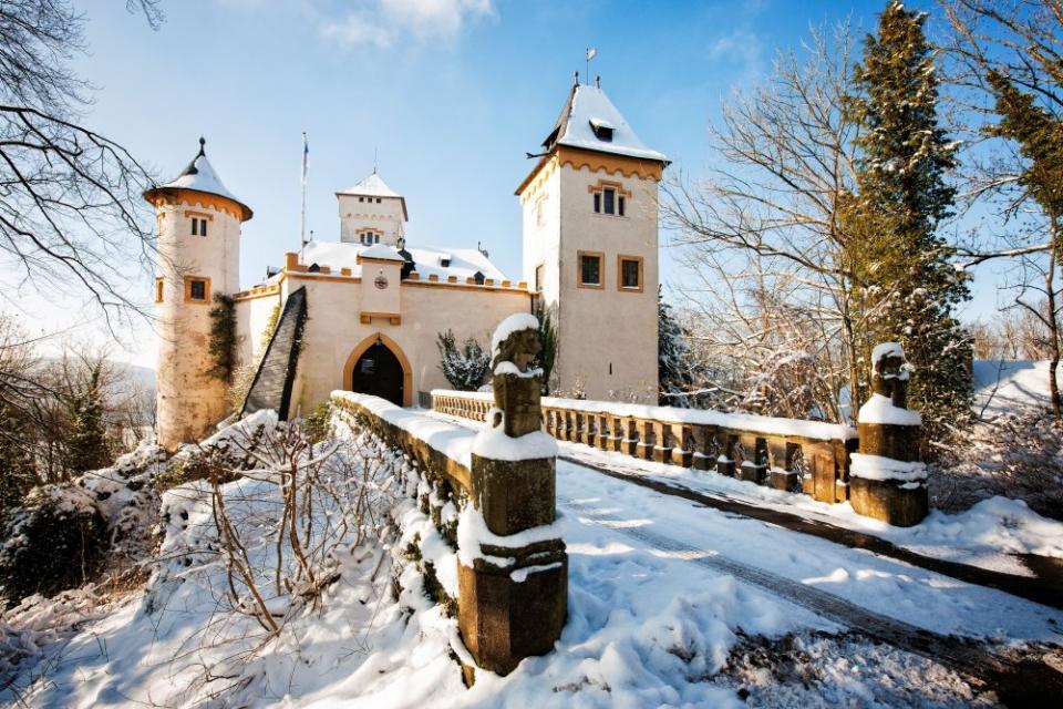Eine schneebedeckte Brücke führt hinüber zum weiß-gelben Schloss mit drei Türmen.