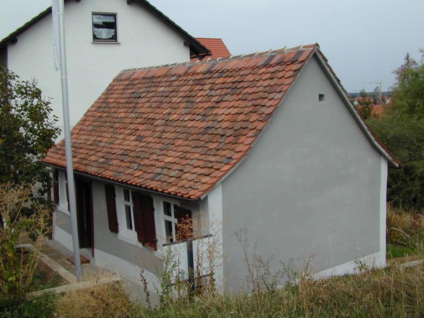 Ein kleines, schlichtes graues Haus mit drei kleinen Fenstern und einer Tür. An den Fenstern sind Fensterläden angebracht.
                 title=