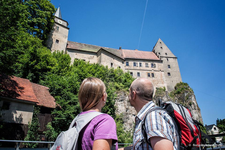 Zwei Wanderer im Vordergrund blicken nach oben zum Schloss, welches auf einem Fels thront.