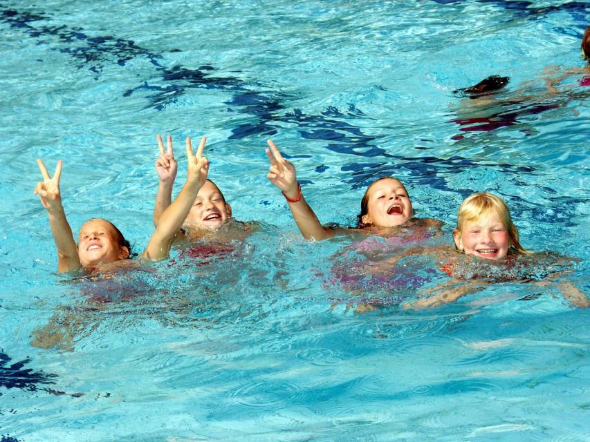 Vier Kinder im blauen Becken lachen und strecken die Hände nach oben.