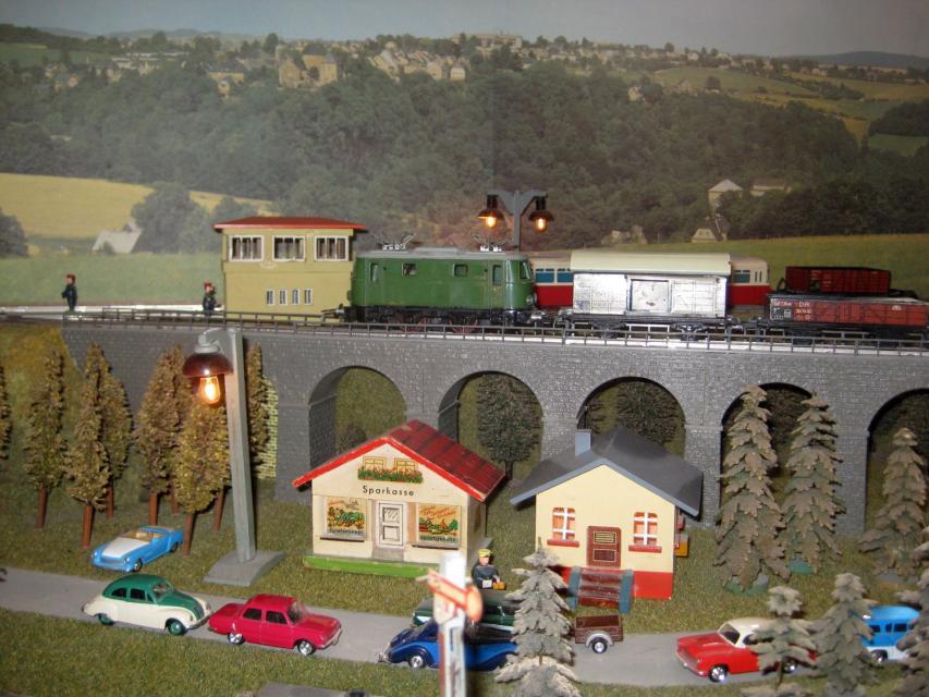 Über eine steinerne Brücke fährt eine Lok mit Anhängern. Unter der Brücke stehen Häuser und auf einer Straße Modellautos.