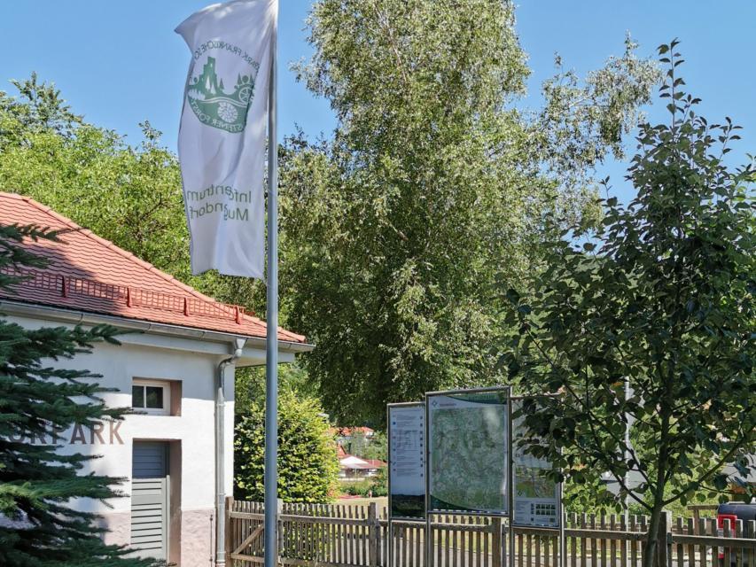 An einem Hauseck steht eine Fahnenmast mit weißer Fahne. Rechts daneben schließt sich ein Zaun an. Hinter dem Zaun stehen drei Infotafeln für Wanderer.