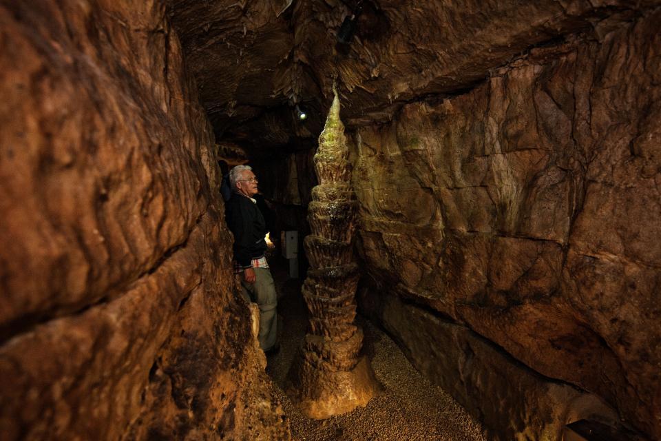In einem schmalen Gang in der Höhle, der links und rechts von Felswänden begrenzt ist, erhebt sich ein mächtiger Tropfstein. An dem Tropfstein steht ein Mann, der diesen bewundert.