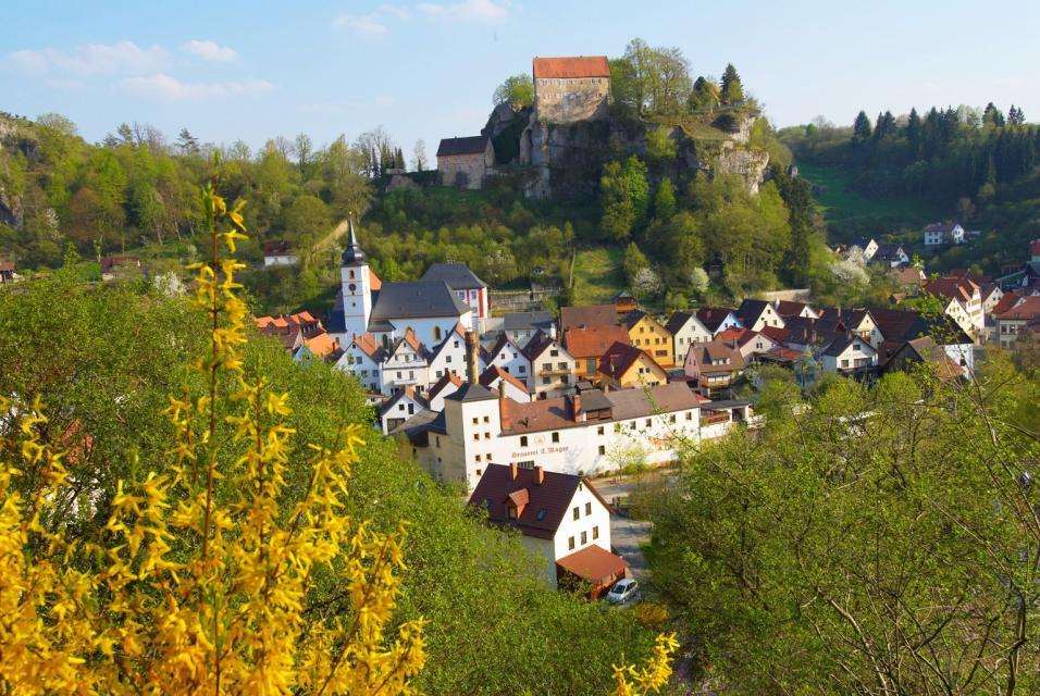 Ortsansicht mit blühender Umrahmung von gelben Sträuchern und grünen Bäumen. Auffällig ist neben der Burg die Kirche Pottensteins und die Brauereigaststätte Mager.