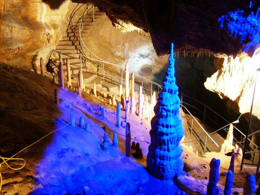 Ein einem großen Höhlenssal erhebt sich blau-violett angestrahlt, ein massiver Tropfstein. Im Hintergrund führen Treppenstufen mit Geländer durch die Höhle.