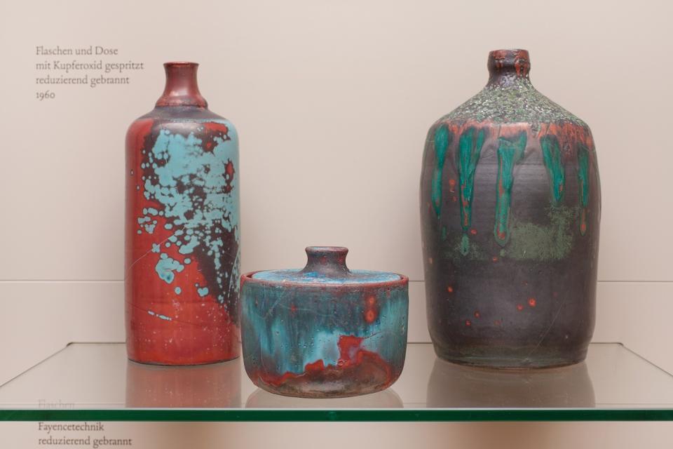 Auf einem Glas stehen drei Töpfereiartikel. Eine rot-blau gefleckte Flasche. Ein kleiner, rot-blau gefleckter Topf mit Deckel und eine bauchige, grün-graue Flasche.