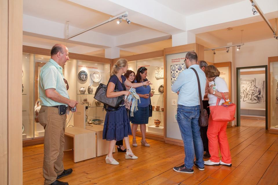 In einem Ausstellungsraum stehen sechs Besucher, die sich von einem Führer die ausgestellten Gegenstände erklären lassen.