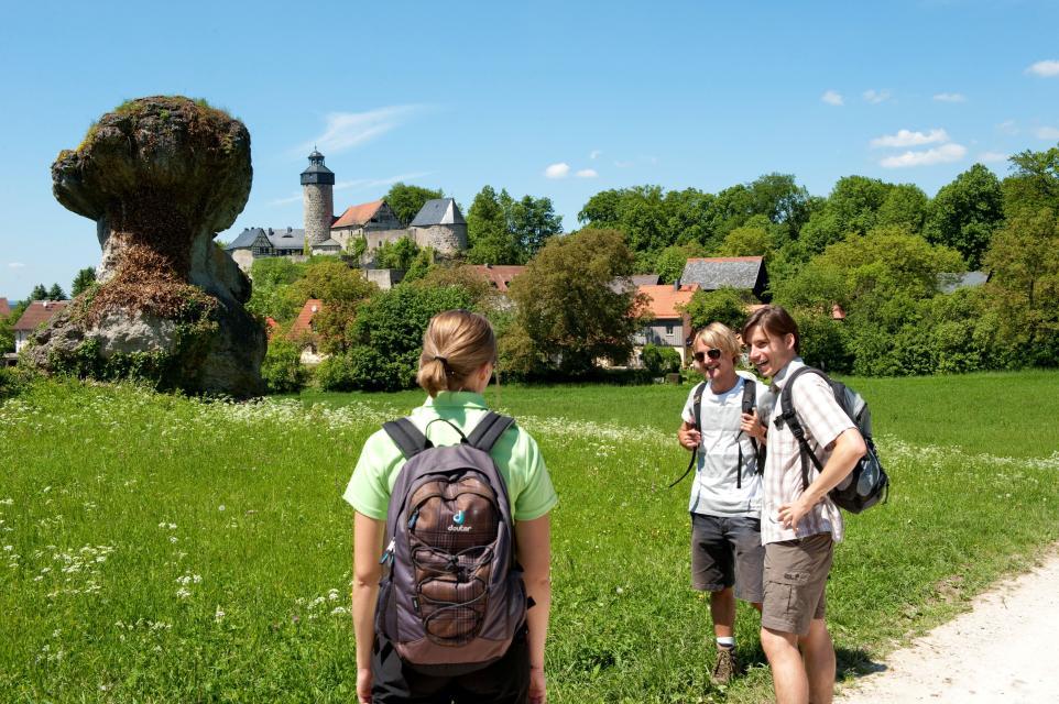 Drei Wanderer unterhalten sich fröhlich an einer blühenden Wiese, auf welchem ein Fels in die Höhe ragt. Der Ort Sanspareil mit der Burganlage Zwernitz ist von Weitem zu erkennen.