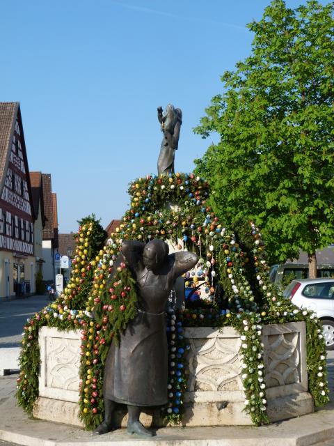 Vor dem steinernen Brunnen steht eine bronzene Frauenfigur. Oben, mittig auf dem Brunnen ist eine weitere Figur zu sehen. Girlanden aus Nadelzweigen und Ostereiern ranken sich nach oben.