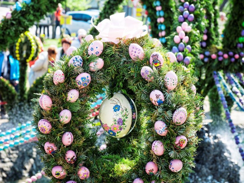 Im Zentrum des Bildes ist ein eiförmiges Grüngesteck, an dessen Rand rosafarbene Eier stecken. In der Mitte hängt eine größeres, mit Blumen handbemaltes Ei.