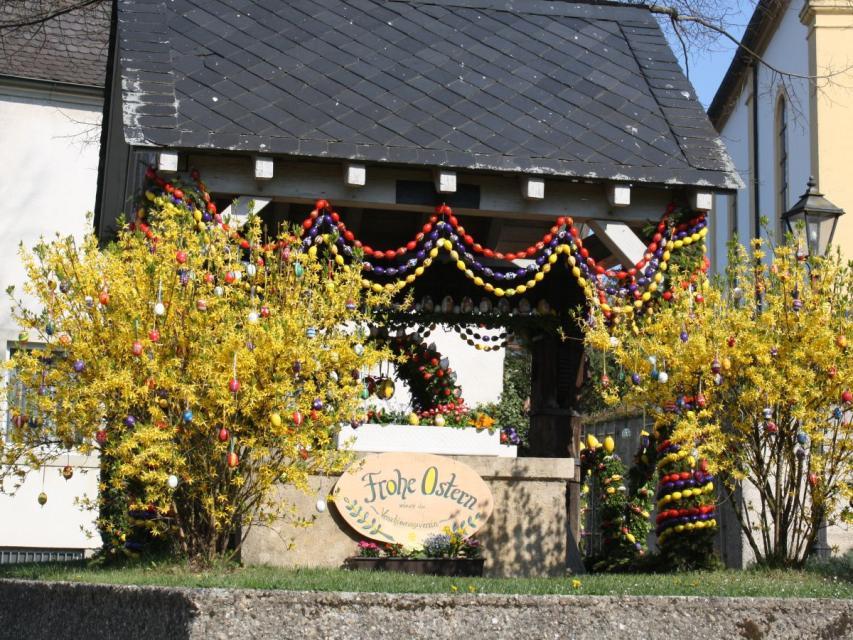 Am Brunnenhäuschen mit schwarzen Schiefer hängen drei Ketten mit roten, blauen und gelben Ostereiern.
