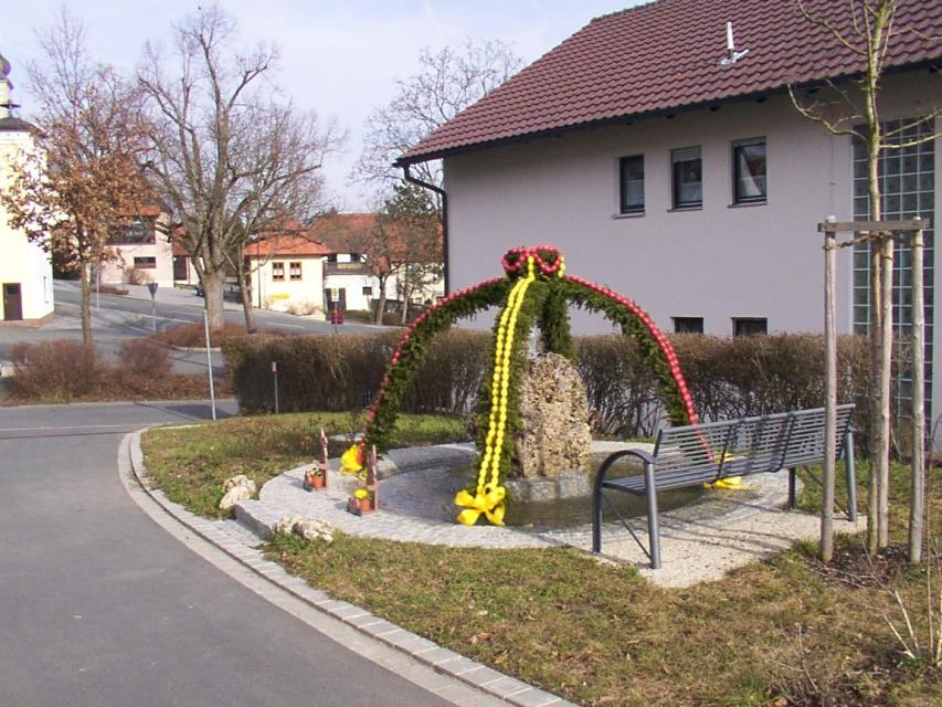 Der Osterbrunnen mit roten und gelben Eiern steht zwischen der Straße und einem Haus.