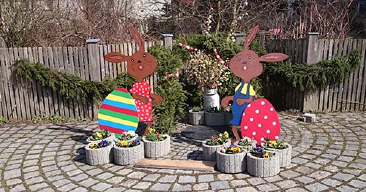 An einem Brunnen, der vor einem Holzzaun steht, an dem Girlanden aus Nadelzweigen hängen, stehen zwei Osterhasenfiguren mit großen bunten Eiern.