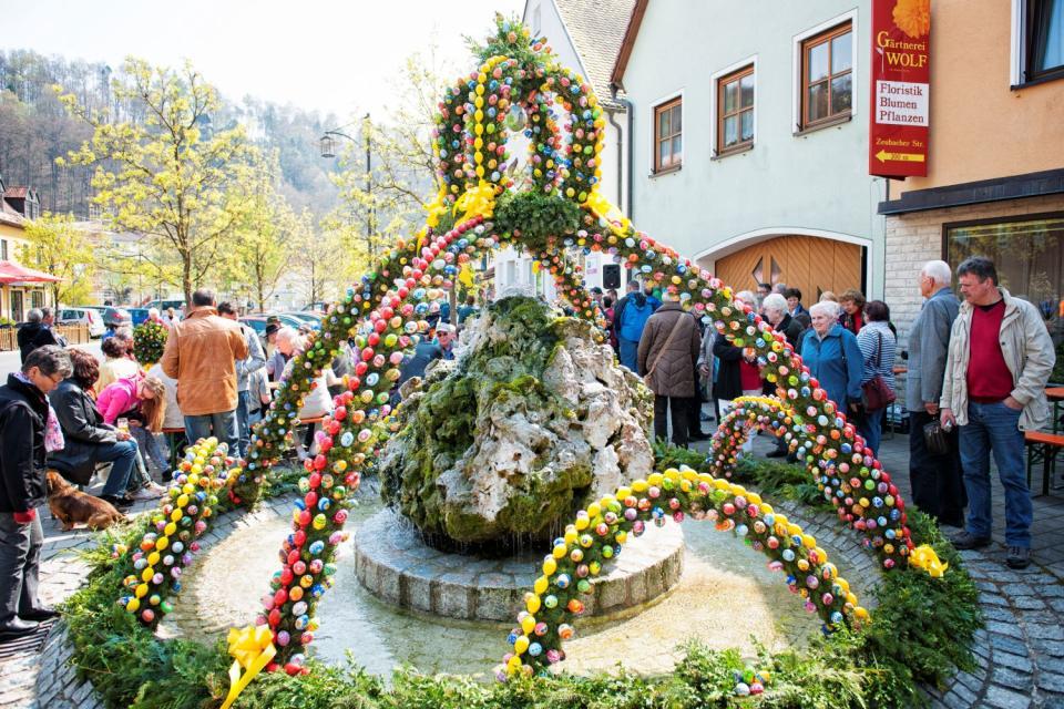 Besucher bewundern den Osterbrunnen, in dessen Mitte ein naturgewachsener Stein steht. Darüber erhebt sich eine Krone aus Nadelzweigen mit bunten Ostereiern.
