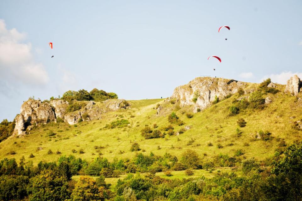 Über einem Hügel, aus dem einzelne Felspartien blitzen, schweben drei Drachenflieger in der Luft.