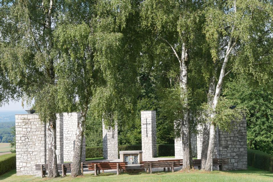 Blick auf das halbrund Ehrendenkmal mit den weißen Steinsäulen unter grünenden Laubbäumen.