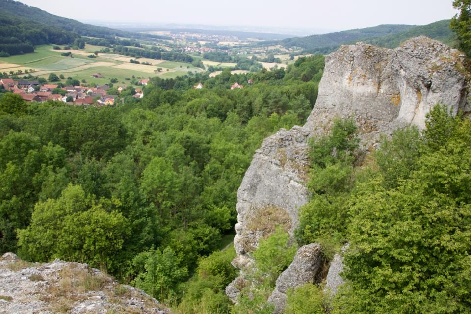 Blick vom Aussichtspunkt in das weite und sanfte grüne Tal. Im Vordergrund rechts erhebt sich ein mächtiger Fels.