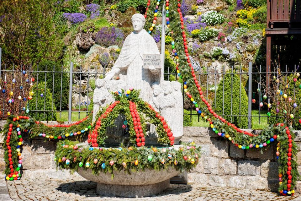 Vor einer christlichen, weißen Statue steht ein runder Osterbrunnen, über den kronenartig Girlanden führen, die mit bunten Eiern verziert sind.
                 title=