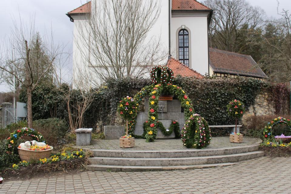 Vor einer Kirche, auf einem kleinen Platz, zu dem zwei Treppenstufen führen, steht der Betonbrunnen. Über dem Brunnen steht eine Krone aus grünen Girlanden mit bunten Eiern.