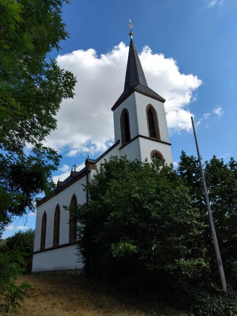 Die weiße Kirche ist geprägt von auf der Sichtseite drei bodentiefen Fenstern. Auch der Glockenturm ist mit solch Fenstern ausgestattet. Geschmückt wird sie durch antrazit gedeckte Dächer.
                 title=