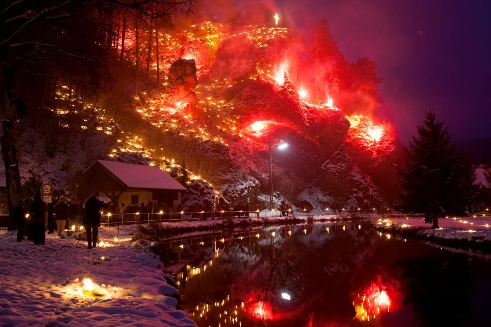 Ein Fluss fließt ruhig zwischen schneebedeckten Wiesen. Im Hintergrund erhebt sich ein Berg, auf dem unzählig viele kleine Feuer brennen und hell leuchten.