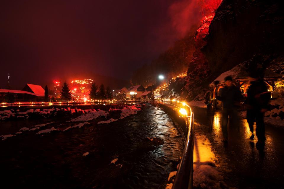 Besucher laufen bei Nacht am Flussufer entlang. Im Hintergrund sind brennende Feuerstellen zu erkennen.