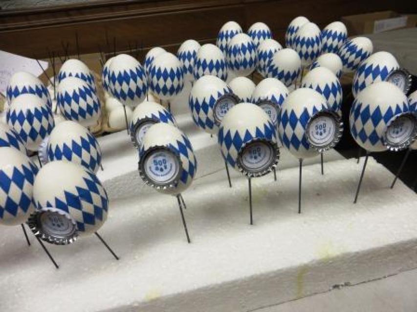 Eine Vielzahl an weißen Eiern, auf denen in Blau die Bayernraute gemalt ist. An jedem Ei klebt ein Kronkorken mit eine Signet im Inneren.